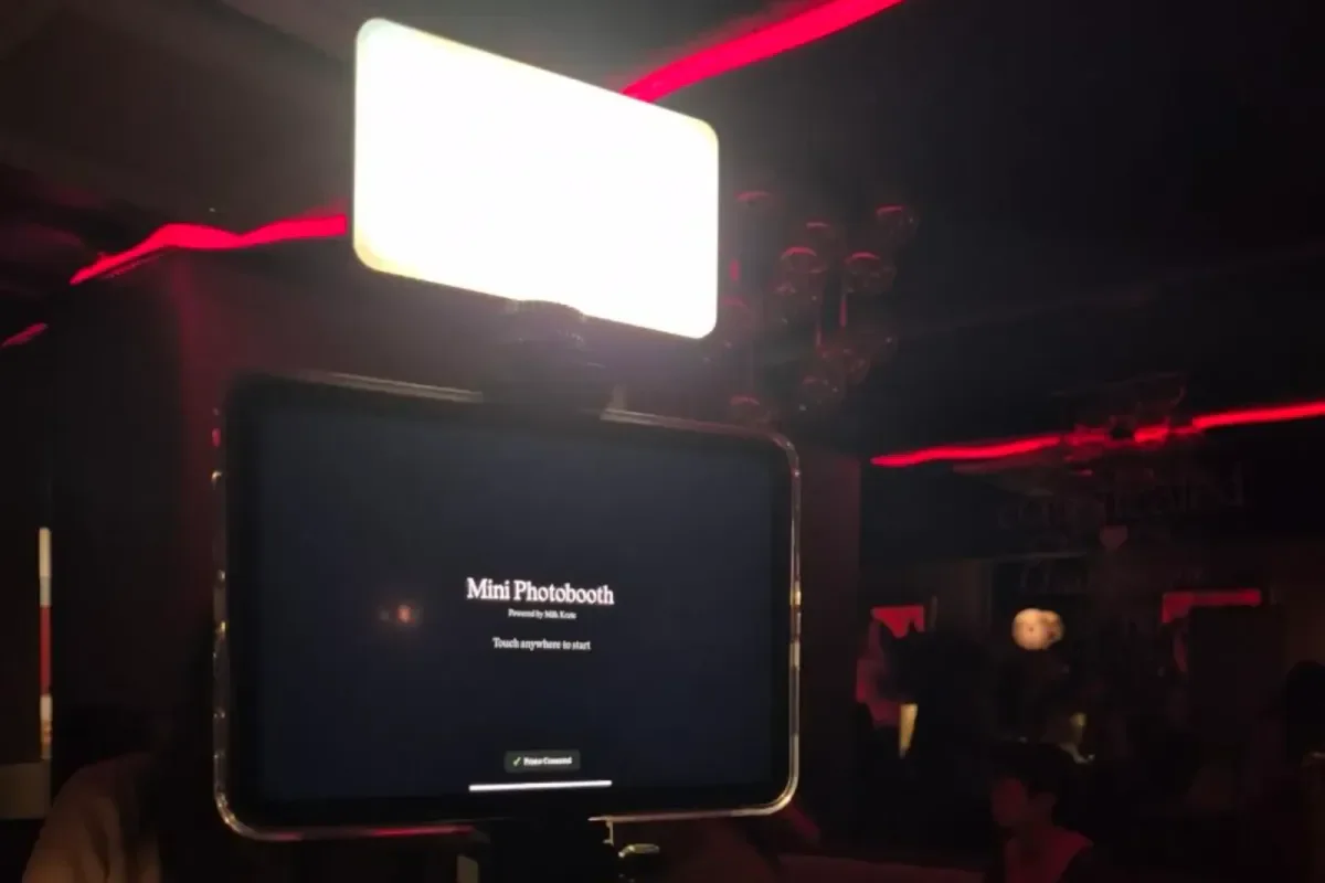 The app's start screen on a tablet, "Mini Photobooth: Touch anywhere to start", lit by a light panel in a dark venue.