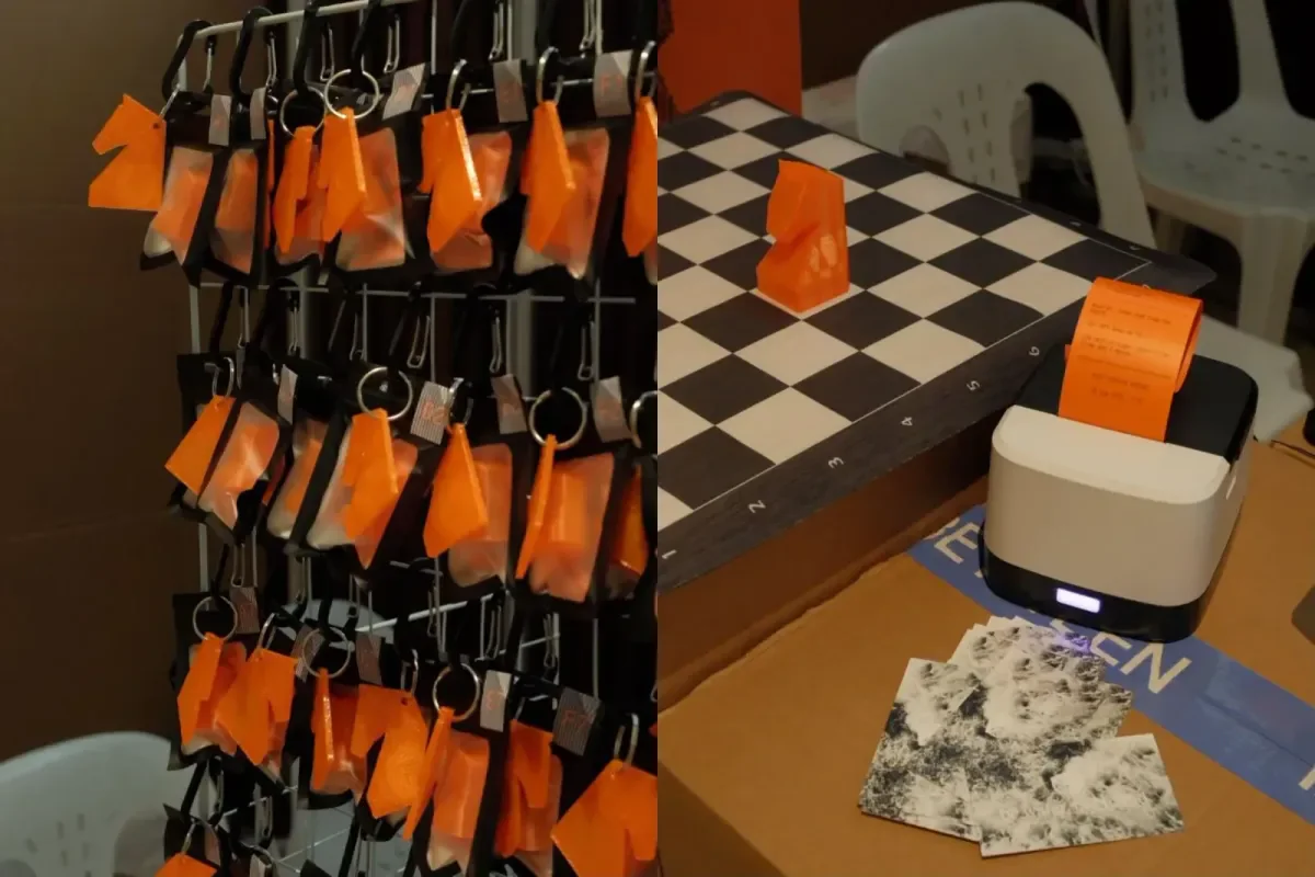 The booth at the festival: a rack of orange 3D-printed horse keychains beside a chessboard with an orange knight, and a receipt printer feeding out an orange receipt.